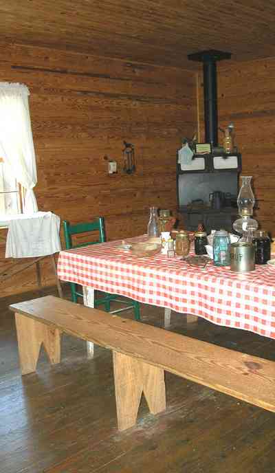 Cantonment:-Roy-Hyatt-Environmental-Center-Dog-Trot-House_17.jpg:  iron bed, quilt, ragdoll, bedroom, wooden walls, wood floor, bench, lantern, wood burning stove, ladder-back chair, ironing board, red checked tablecloth, oil lantern, rocking chair