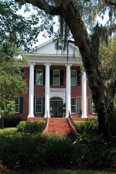 Gulf-Breeze:-506-Kenilworth-Drive_01.jpg:  red brick mansion, white columns, leaded glass door, mailbox, impatiens, brick wall, spanish moss, oak trees, staircase, wrought-iron bannisters, doric columns, pediment, shutters, porch