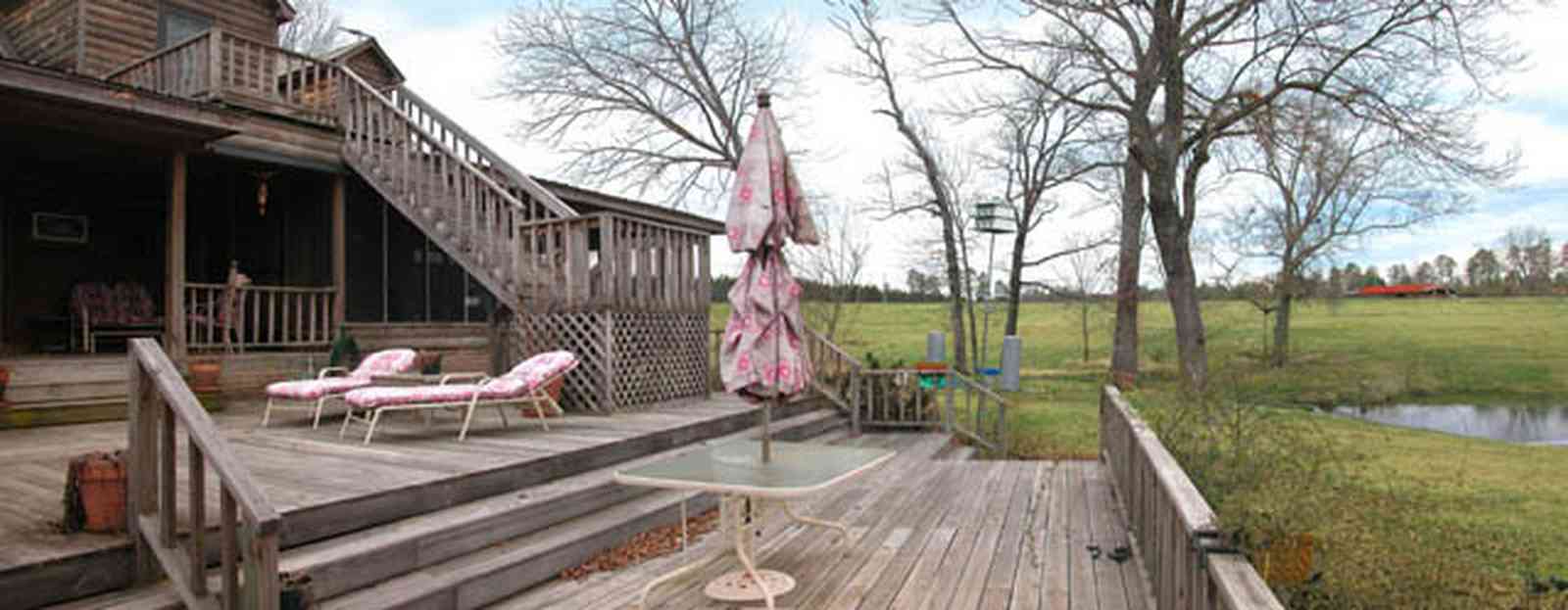 Milton:-Floyd-Farm_05a.jpg:  porch, deck, fireplace, log cabin, hills, valley, pond, oak trees, sofa, chaise lounge, umbrella table, lake